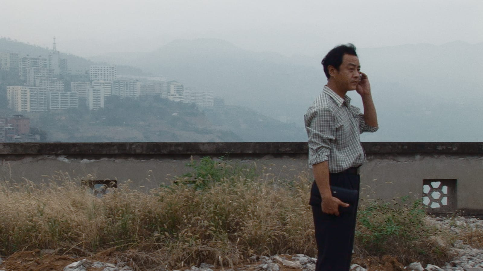 still from Caught by the Tides - a man takes a phone call on a roof, with mountains and apartment buildings in the distant background